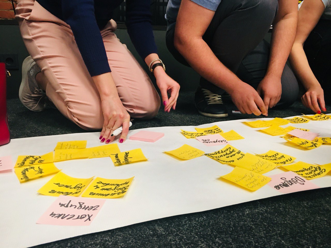 A group of people are sitting on the floor, taking notes as they look at the stickers glued to a large piece of paper